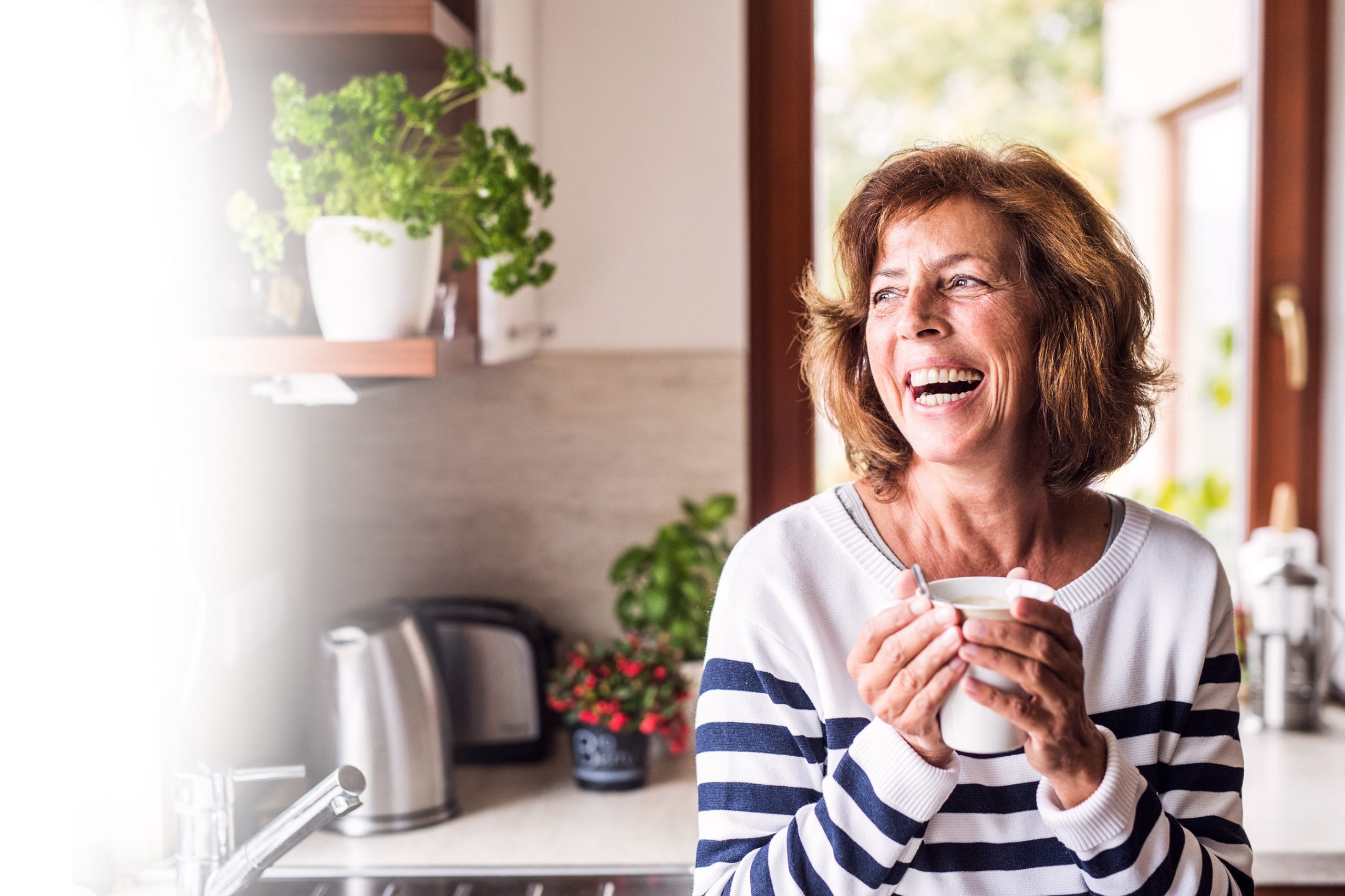 Older Woman looking out a window, smiling, and holding a cup of coffee